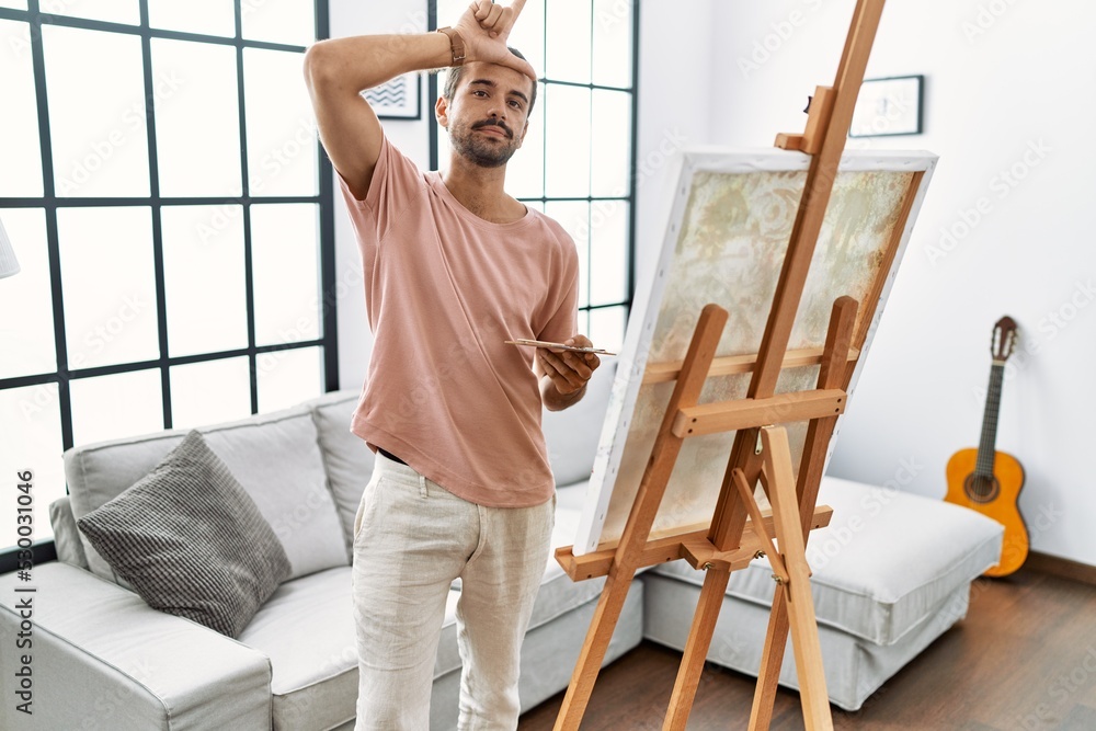 Young hispanic man with beard painting on canvas at home making fun of people with fingers on forehead doing loser gesture mocking and insulting.
