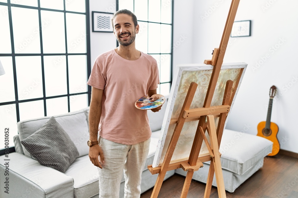 Young hispanic man with beard painting on canvas at home winking looking at the camera with sexy expression, cheerful and happy face.