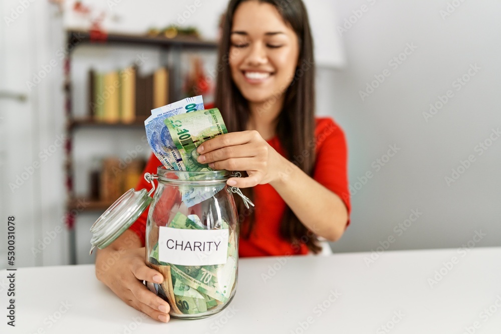 Young latin woman holding charity jar with rands banknotes sitting by christmas decor at home Young latin woman holding charity jar with rands banknotes sitting by christmas decor at home