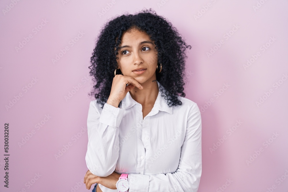 Hispanic woman with curly hair standing over pink background with hand on chin thinking about question, pensive expression. smiling with thoughtful face. doubt concept. Hispanic woman with curly hair standing over pink background with hand on chin thinking about question, pensive expression. smiling with thoughtful face. doubt concept.