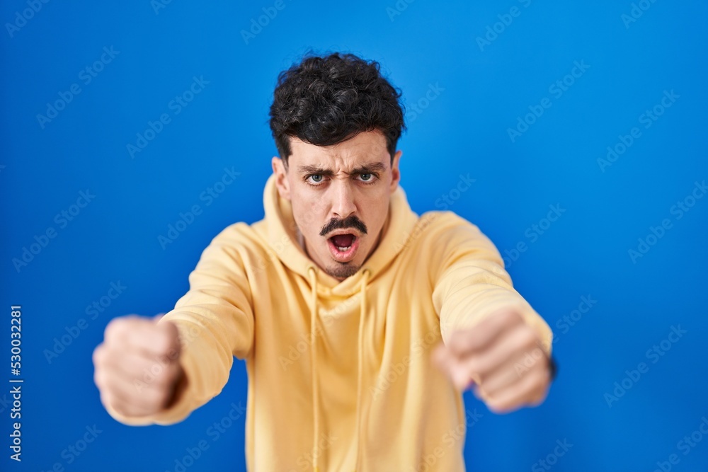 Hispanic man standing over blue background angry and mad raising fists frustrated and furious while shouting with anger. rage and aggressive concept. Hispanic man standing over blue background angry and mad raising fists frustrated and furious while shouting with anger. rage and aggressive concept.