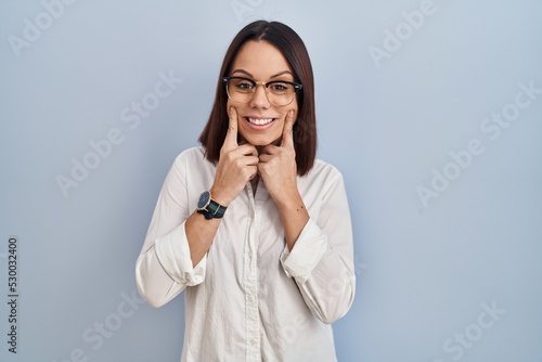 Young hispanic woman standing over white background smiling with open mouth, fingers pointing and forcing cheerful smile