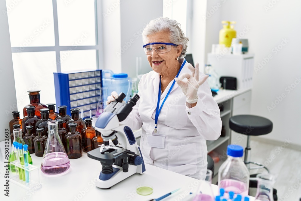 Senior woman with grey hair working at scientist laboratory doing ok sign with fingers, smiling friendly gesturing excellent symbol Senior woman with grey hair working at scientist laboratory doing ok sign with fingers, smiling friendly gesturing excellent symbol
