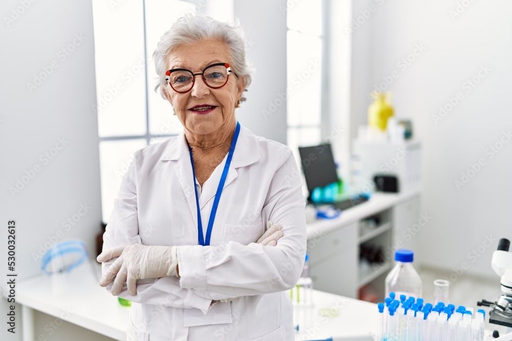 Senior grey-haired woman wearing scientist uniform standing with arms crossed gesture at laboratory