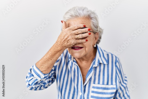 Senior woman with grey hair standing over white background smiling and laughing with hand on face covering eyes for surprise. blind concept.