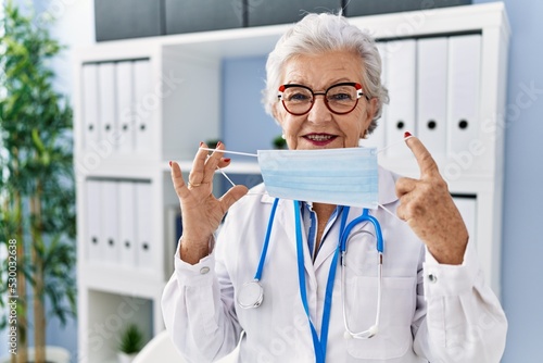 Senior grey-haired woman wearing doctor uniform and medical mask at clinic