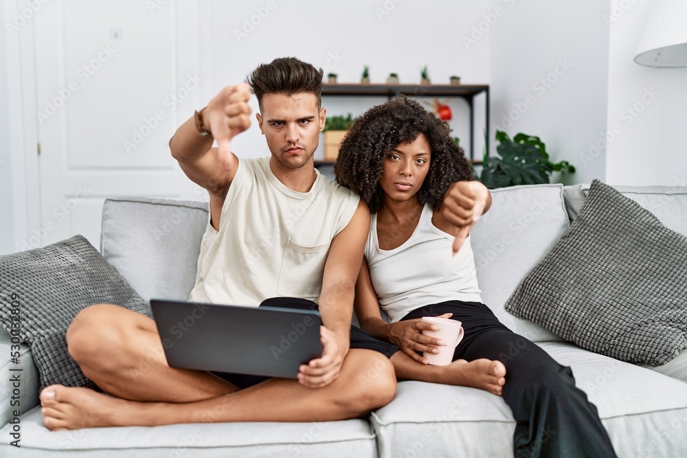 Young interracial couple using laptop at home sitting on the sofa looking unhappy and angry showing rejection and negative with thumbs down gesture. bad expression. Young interracial couple using laptop at home sitting on the sofa looking unhappy and angry showing rejection and negative with thumbs down gesture. bad expression.