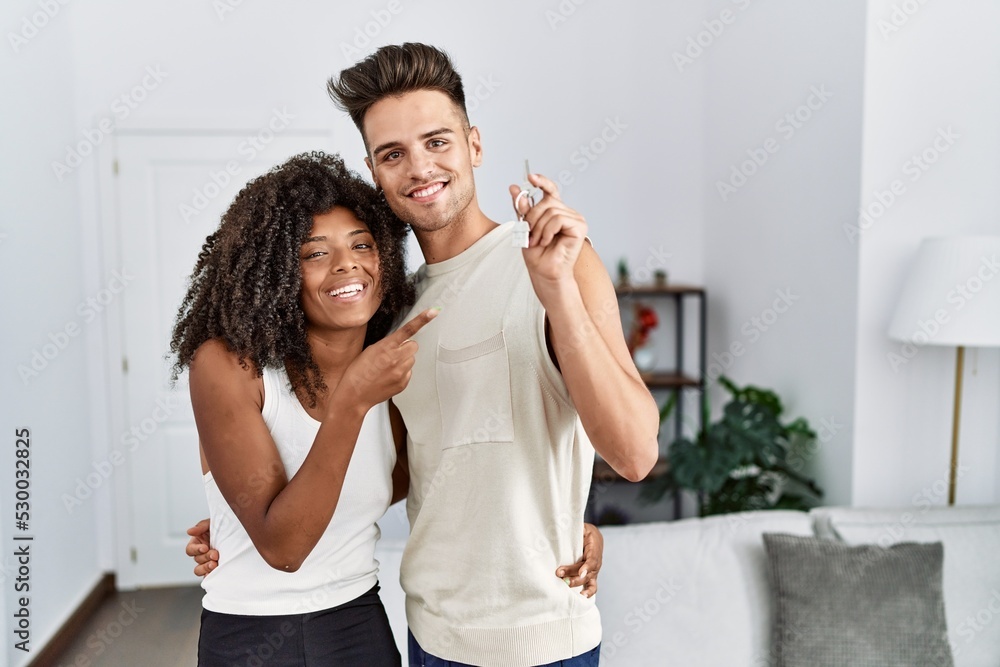 Obraz premium Young interracial couple holding keys of new home smiling happy pointing with hand and finger