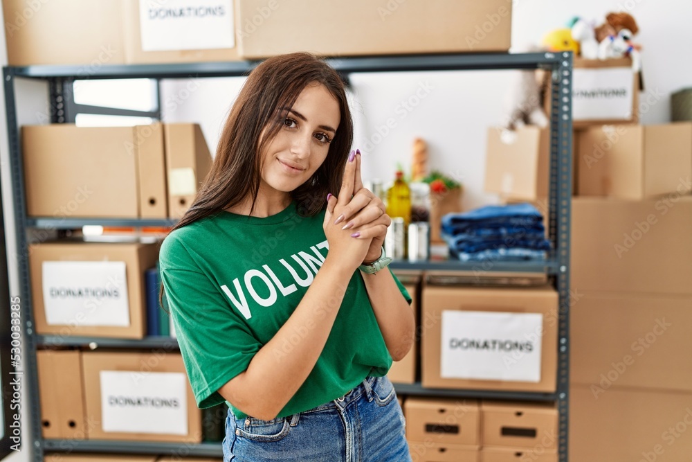 Young brunette woman wearing volunteer t shirt at donations stand holding symbolic gun with hand gesture, playing killing shooting weapons, angry face Young brunette woman wearing volunteer t shirt at donations stand holding symbolic gun with hand gesture, playing killing shooting weapons, angry face