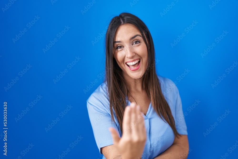 Young brunette woman standing over blue background beckoning come here gesture with hand inviting welcoming happy and smiling Young brunette woman standing over blue background beckoning come here gesture with hand inviting welcoming happy and smiling