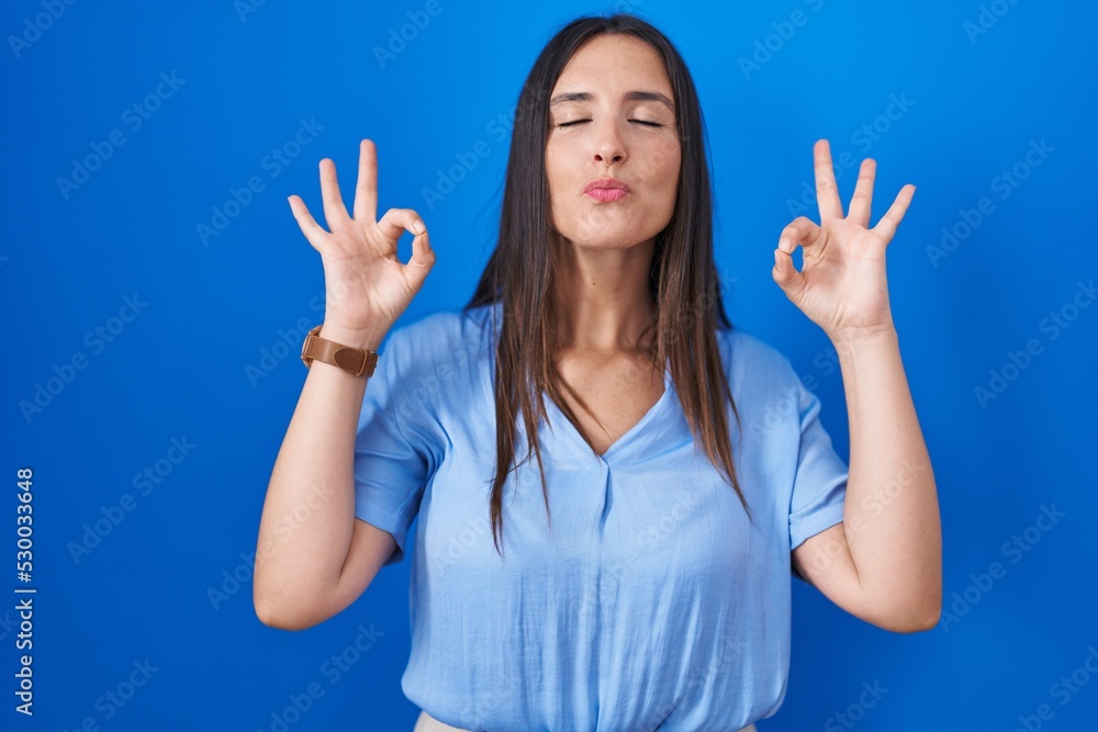 Young brunette woman standing over blue background relaxed and smiling with eyes closed doing meditation gesture with fingers. yoga concept. Young brunette woman standing over blue background relaxed and smiling with eyes closed doing meditation gesture with fingers. yoga concept.
