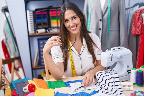 Young beautiful hispanic woman tailor smiling confident sitting on table at clothing factory