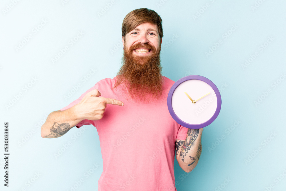 Young caucasian man holding a clock isolated on blue background person pointing by hand to a shirt copy space, proud and confident Young caucasian man holding a clock isolated on blue background person pointing by hand to a shirt copy space, proud and confident