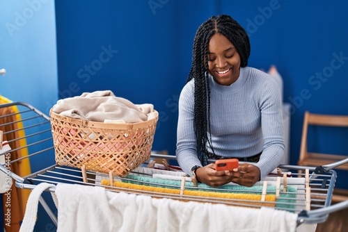 African american woman using smartphone hanging clothes on clothesline at laundry room