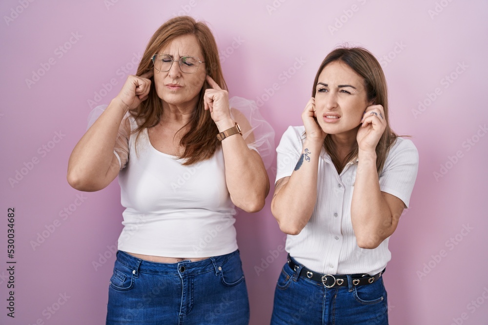 Hispanic mother and daughter wearing casual white t shirt over pink background covering ears with fingers with annoyed expression for the noise of loud music. deaf concept. Hispanic mother and daughter wearing casual white t shirt over pink background covering ears with fingers with annoyed expression for the noise of loud music. deaf concept.