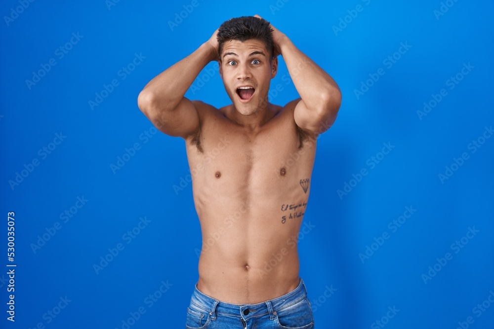 Fototapeta premium Young hispanic man standing shirtless over blue background crazy and scared with hands on head, afraid and surprised of shock with open mouth