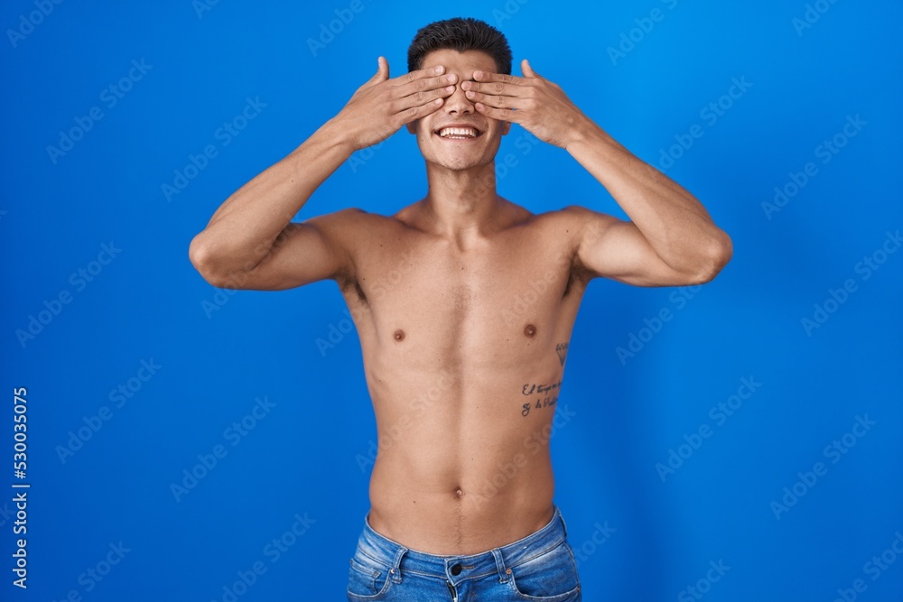Fototapeta premium Young hispanic man standing shirtless over blue background covering eyes with hands smiling cheerful and funny. blind concept.
