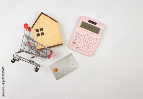 flat layout of wooden house model in shopping trolley, pink calculator and credit card on white background, home purchase calculation concept.
