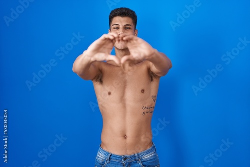 Young hispanic man standing shirtless over blue background smiling in love doing heart symbol shape with hands. romantic concept.