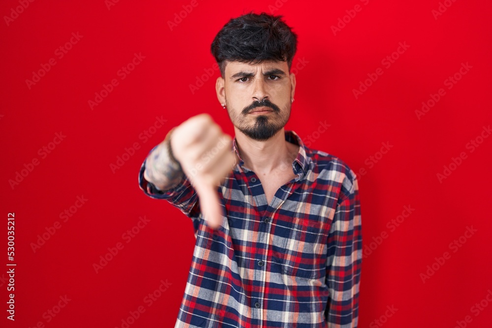 Fototapeta premium Young hispanic man with beard standing over red background looking unhappy and angry showing rejection and negative with thumbs down gesture. bad expression.
