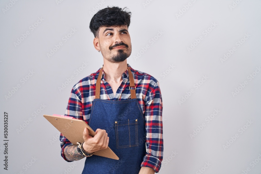 Young hispanic man with beard wearing waiter apron holding clipboard smiling looking to the side and staring away thinking.