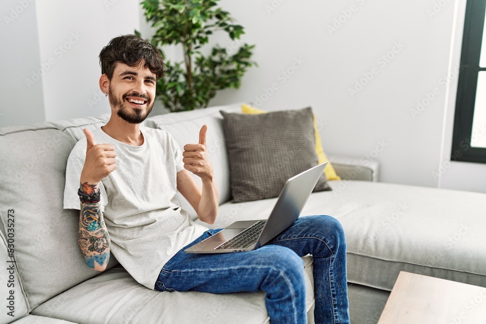 Hispanic man with beard sitting on the sofa success sign doing positive gesture with hand, thumbs up smiling and happy. cheerful expression and winner gesture.