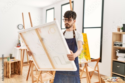 Young hispanic artist man concentrate holding canvas at art studio.