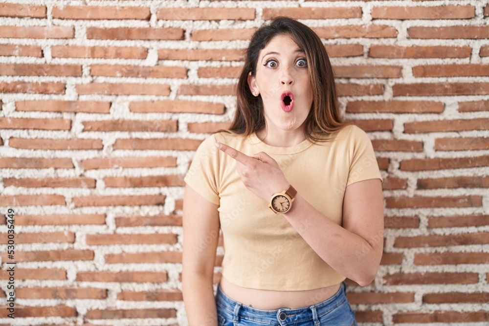 Young brunette woman standing over bricks wall surprised pointing with finger to the side, open mouth amazed expression. Young brunette woman standing over bricks wall surprised pointing with finger to the side, open mouth amazed expression.