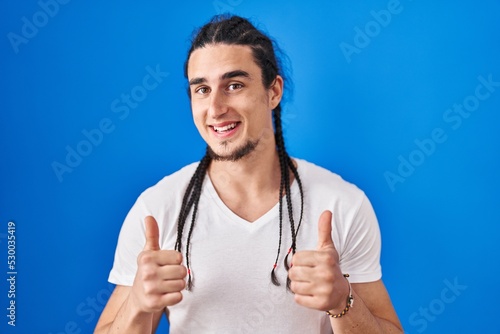 Hispanic man with long hair standing over blue background success sign doing positive gesture with hand, thumbs up smiling and happy. cheerful expression and winner gesture.