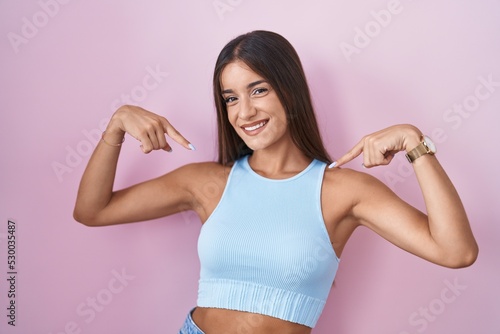Young brunette woman standing over pink background looking confident with smile on face, pointing oneself with fingers proud and happy.