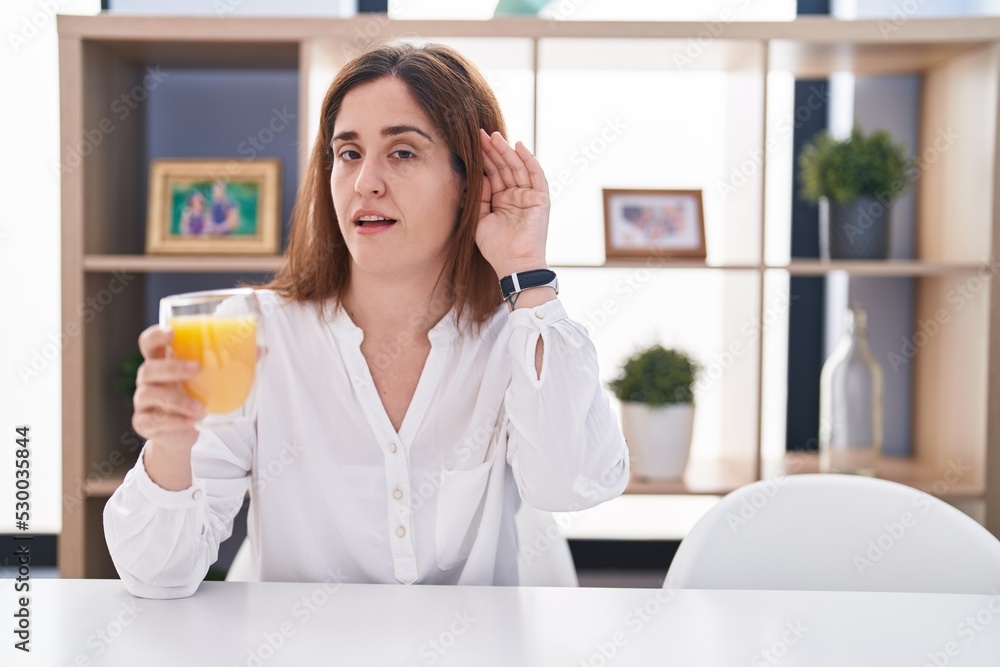 Brunette woman drinking glass of orange juice smiling with hand over ear listening an hearing to rumor or gossip. deafness concept. Brunette woman drinking glass of orange juice smiling with hand over ear listening an hearing to rumor or gossip. deafness concept.