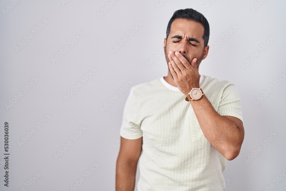 Hispanic man with beard standing over isolated background bored yawning tired covering mouth with hand. restless and sleepiness. Hispanic man with beard standing over isolated background bored yawning tired covering mouth with hand. restless and sleepiness.