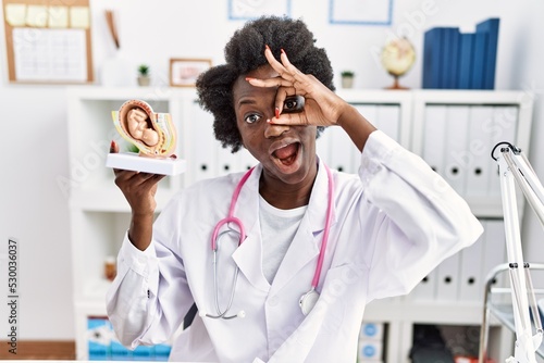 African doctor woman holding anatomical model of female uterus with fetus smiling happy doing ok sign with hand on eye looking through fingers