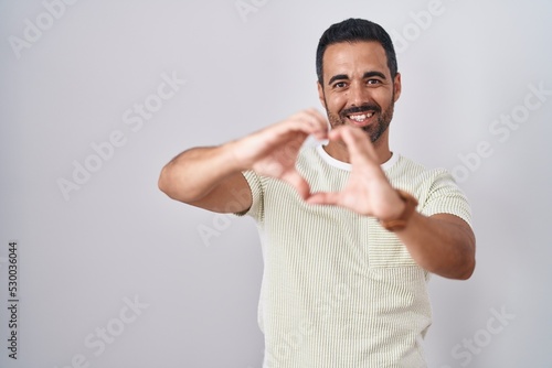 Hispanic man with beard standing over isolated background smiling in love doing heart symbol shape with hands. romantic concept.