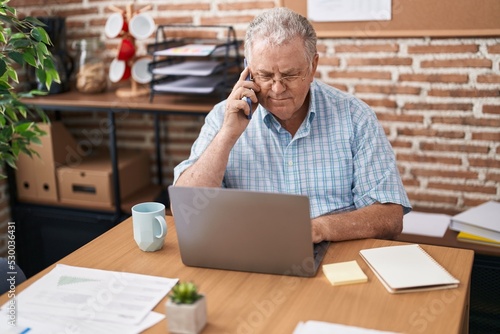 Middle age grey-haired man business worker using laptop talking on smartphone at office