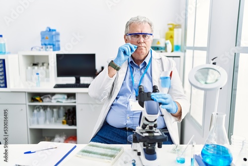Senior caucasian man working at scientist laboratory mouth and lips shut as zip with fingers. secret and silent, taboo talking