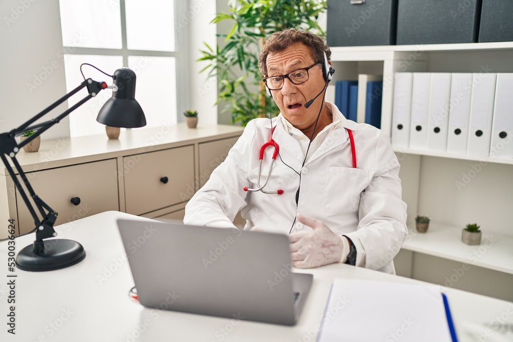 Obraz premium Senior doctor man working on online appointment in shock face, looking skeptical and sarcastic, surprised with open mouth