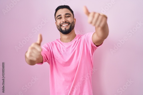 Hispanic young man standing over pink background approving doing positive gesture with hand, thumbs up smiling and happy for success. winner gesture.
