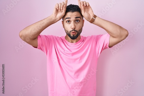 Hispanic young man standing over pink background doing funny gesture with finger over head as bull horns