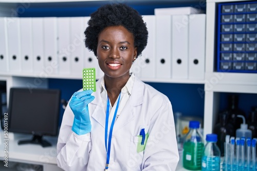 African young woman working at scientist laboratory holding birth control pills looking positive and happy standing and smiling with a confident smile showing teeth photo