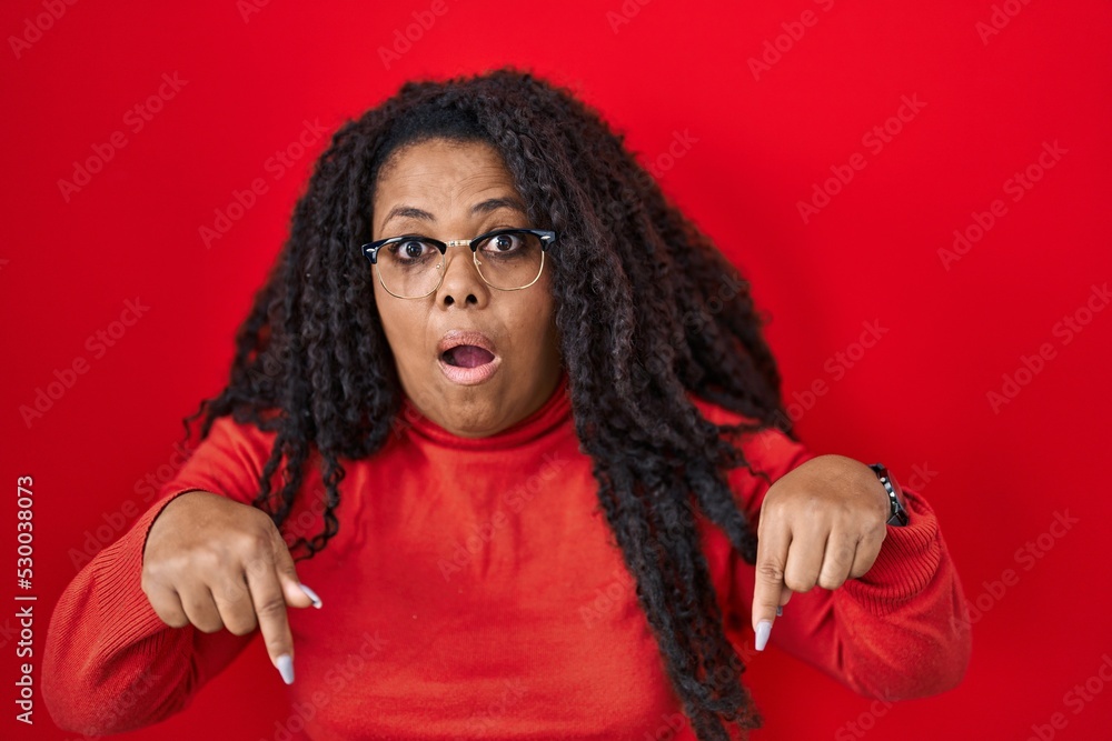 Plus size hispanic woman standing over red background pointing down with fingers showing advertisement, surprised face and open mouth Plus size hispanic woman standing over red background pointing down with fingers showing advertisement, surprised face and open mouth