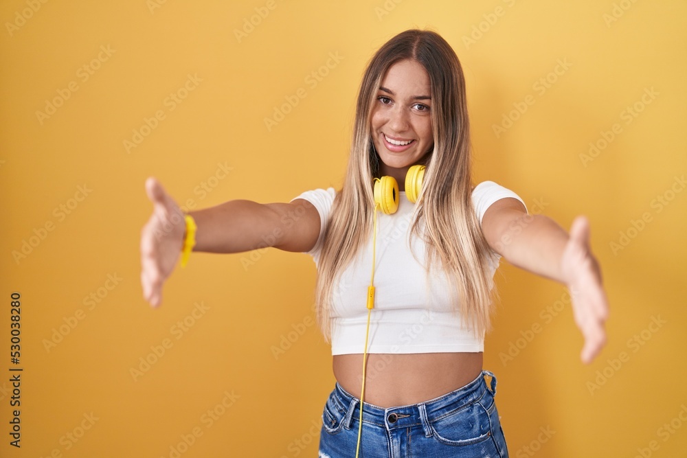 Young blonde woman standing over yellow background wearing headphones looking at the camera smiling with open arms for hug. cheerful expression embracing happiness. Young blonde woman standing over yellow background wearing headphones looking at the camera smiling with open arms for hug. cheerful expression embracing happiness.