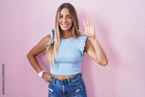 Young blonde woman standing over pink background waiving saying hello happy and smiling  friendly welcome gesture