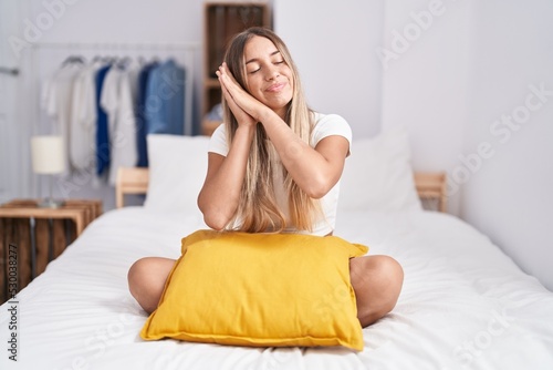 Young blonde woman sitting on the bed with pillow at home sleeping tired dreaming and posing with hands together while smiling with closed eyes.
