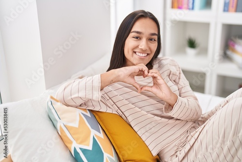 Young beautiful hispanic woman doing heart gesture sitting on bed at bedroom