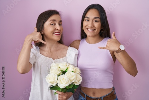 Hispanic mother and daughter holding bouquet of white flowers smiling doing phone gesture with hand and fingers like talking on the telephone. communicating concepts.