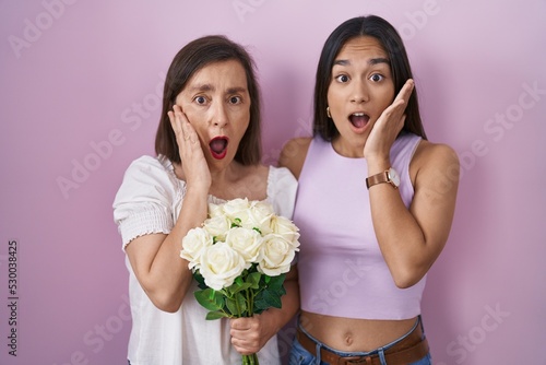 Hispanic mother and daughter holding bouquet of white flowers afraid and shocked, surprise and amazed expression with hands on face