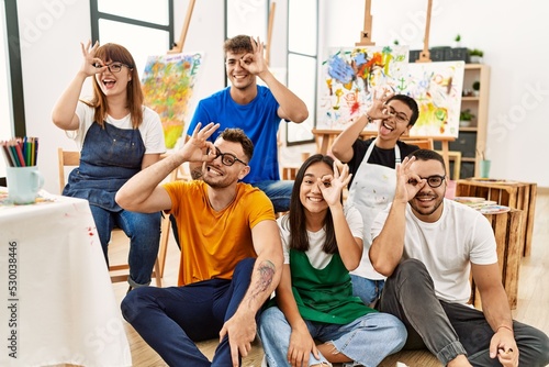 Group of people sitting at art studio smiling happy doing ok sign with hand on eye looking through fingers