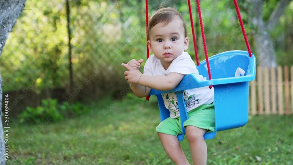Sweet Caucasian baby boy with bare feet sits in a swing. Cute child having good time outdoors in summer. Blurred nature backdrop. Sweet Caucasian baby boy with bare feet sits in a swing. Cute child having good time outdoors in summer. Blurred nature backdrop.