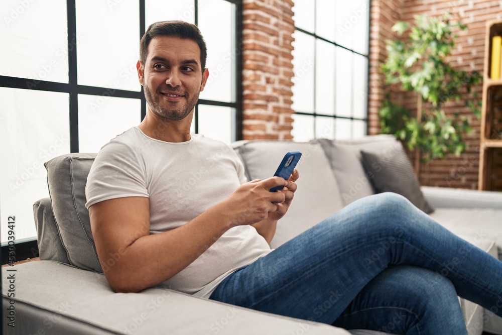 Obraz premium Young hispanic man using smartphone sitting on sofa at home